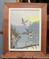 Framed painting showing mallard ducks flying over a lake with trees and a fallen branch in the foreground.