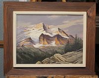 Framed oil painting showing a snowy Rocky Mountain scene with pine trees and rocks under natural lighting.