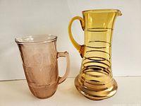 Front view of the two vintage glass jugs showing amber jug with black stripes and light pinkish jug with etched floral pattern side by side.