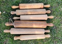 Photo of six vintage wooden rolling pins laid out on grass. Each rolling pin has two handles, varying in wood tone and size.