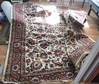 Full view of the large rug laid partially rolled, showing floral and geometric pattern in red, cream, gold, and black colors with fringed edges.