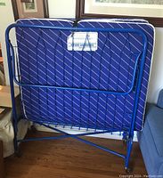 Fold-up metal frame cot with blue wire mesh, blue mattress with diagonal white stripes folded upright, casters visible.
