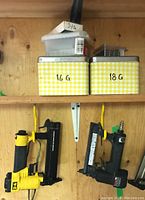 Two pneumatic brad nailers hanging on hooks below two metal tins of 16 gauge and 18 gauge nails, along with a small plastic container of 3/4 nails on a shelf.