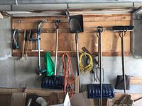 Wide view of all garden tools hanging on wooden wall with extension cords coiled below and cardboard boxes on floor