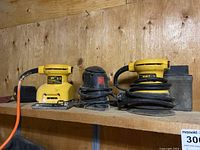 Three palm sanders on wooden shelf: two yellow DeWalt palm sanders and one black Jobmate sander between them. DeWalt models have power cords wrapped around them.