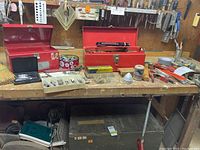 Photo of workbench with two open red metal toolboxes containing various hand tools, a plastic organizer with screws, and assorted items on the bench surface. Workbench is wooden with multiple small items scattered.