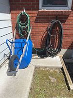 Photo showing the blue steel wheelbarrow with wooden handles, single black rubber wheel, and two garden hoses (green and black) coiled on wall-mounted holders which are not included.