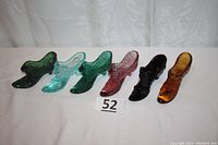 Six Fenton art glass slippers in a row: dark green, light aqua, emerald green, pink, black, and amber colors. Five have cat heads and floral designs; one amber slipper has no cat head.