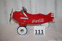 Side profile of red die cast Coca-Cola branded pedal plane showing logo and white wheels.