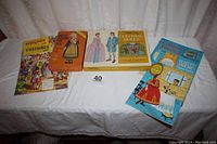 Seven boxed sets of vintage paper dolls arranged on a white surface showing front covers with titles and illustrations of costumes and dolls