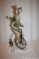 Full view of the cottontail bunny doll sitting on a black high wheel bicycle with a white base, showing the bunny's pose and the broken strings on its hands.