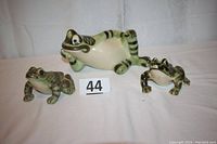 Group photo showing three ceramic frog figurines in cream and green colors, arranged with the largest reclining frog in center and two smaller frogs beside it.