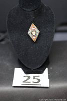 Front view of diamond-shaped colorful micro mosaic brooch on black display stand with identification card below labeled 25.