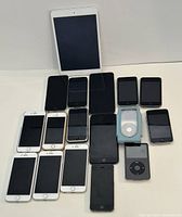 Lot of Apple devices including multiple iPhones, iPads, iPods spread out flat showing front of devices.