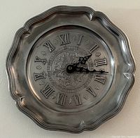 Full frontal photo of the vintage pewter wall clock showing scalloped edges and clock face with Roman numerals.