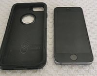 Front view of iPhone SE next to OtterBox case on light fabric surface. Phone screen is off, home button visible.