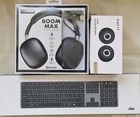 Photo showing front view of Fisher BOOM MAX wireless Bluetooth headphones in black packaging, HAVIT USB stereo speakers packaging, and wired keyboard packaging.
