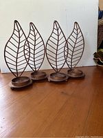 Four copper leaf pattern candleholders shown standing upright on wooden floor against white wall. Each has a wireframe leafback and round base.