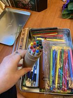 Metal container filled with assorted art supplies including colorful crayons, ballpoint pens, wooden rulers, and paintbrushes.
