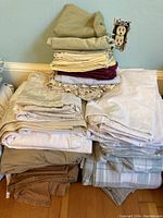 Stacked assorted queen size bed linens including sheets and pillowcases in beige, cream, white, burgundy, floral, and striped patterns on a flat surface.
