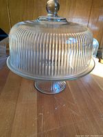 Clear glass cake stand with ribbed pattern and matching lid with knob handle on top shown from above and side angles.