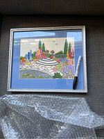 Framed embroidered garden scene picture showing full artwork with silver frame and blue mat, pen for scale.