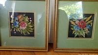 Two framed floral cross stitch hand stitched artworks side by side, with gold-tone frames and green mattes