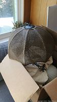 Woven wicker dome-shaped pendant light shade in brown, shown inside cardboard box near window.