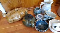 Pottery assortment showing multiple handmade bowls, a teapot, a pitcher, and a large lidded casserole dish on wooden surface.