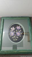 Framed oval tufted moose hair art depicting purple flowers and a hummingbird, signed by Gladys Lavalle.