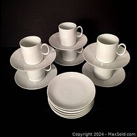 Six white porcelain demi-tasse cups each paired with wide-rimmed saucers and six round side plates, set against black background.