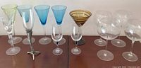 Full view of all 13 stemware glasses arranged on two surfaces showing variety in colors and styles including clear, blue, green, amber, and pewter giraffe stem.