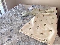 Cal-King bedding set arranged on bed showcasing light blue comforter with beach scenes and multiple folded sheets and pillowcases as well as white cotton blanket with embroidered palm trees