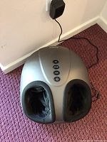 Gray Breo foot massager plugged into wall, showing two black fabric foot openings and power cord.