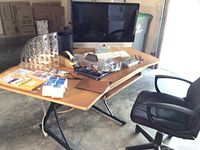 Wide angle view of the handmade wooden desk with various office supplies, a black office chair, and an Apple iMac monitor with keyboard and mouse in a garage setting.