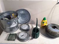 Full view of all items on table including ceramic pot, tin clock, temperature gauge, small bucket with lid, green watering pitcher, bird seed cage, and plant food bottle.