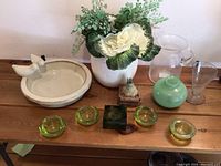 Photo of all items on a wooden surface including green glass ashtrays, white ceramic dish with bird figures, ceramic pot with artificial flowers, green ceramic vase, glass pitcher and vase, and small ceramic planter with artificial succulent.