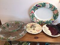 One large clear glass bowl and one very large clear glass platter with leaf decorations. Three hand-painted ceramic platters, including a Christmas-themed platter, a floral platter with handle, and a burgundy/white abstract design platter.