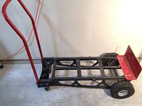 Hand truck showing black plastic frame with red metal handle and red toe plate on concrete floor.