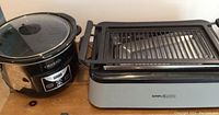 Photo showing black Crock Pot with glass lid missing handle and Simple Living electric grill on a wooden surface.