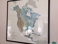 Full view of large framed map of North America on shiny board surface, showing continent and topography.