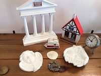 Overall view of the lot showing white plaster column facade, cupid plaque, milk glass hands, camper ornament, house thermometer, and clock on wooden surface.