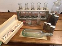 Wooden frame holding five clear glass flower bottles, along with a box of seashells, the ship-in-bottle, small bottle, and metal flasks on a wooden surface.
