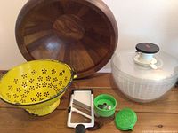 Photo showing large wooden bowl, yellow enamel colander, plastic salad spinner, white slicer, and two green chopper/grater attachments