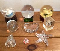 Photo showing three snow globes: one with a swan and missing liquid, one clear globe on green base, and one with two swans. Various glass paperweights including swan, elephant, star and abstract shapes arranged on a wooden surface.