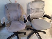 Two gray office chairs side by side showing front view; one with mesh backrest and cushioned seat, the other fully cushioned with padded armrests.