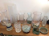 Wide view of eleven glass vases and bowls of different shapes and sizes on a wooden surface, most empty, some filled with glass beads and a few metal flower arranging pieces.