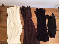 8 vintage ladies gloves displayed on wood surface. 5 pairs creamy white gloves, 1 brown pair, 2 black pairs visible. Variety of lengths including opera and 3/4 length.