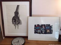 Framed charcoal enlargement of an electric plug beside framed neon photograph and a clock on wooden surface.