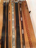 Photo showing six women's belts arranged on a wooden surface. Includes a tan braided leather belt, a dark brown leather belt with metal conchos, a brown faux crocodile belt, a plain black leather belt, a rhinestone and floral decorated belt, and a beaded elastic belt.